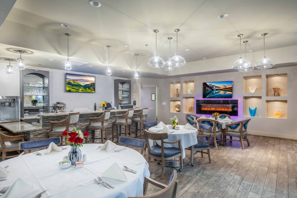 A dining room with tables covered in white table cloth that also feature flower bouquets in the middle of each table. Blue cushioned chairs surround each table, and the room also includes a bar with barstools and colorful vases that sit on the shelves on the wall.