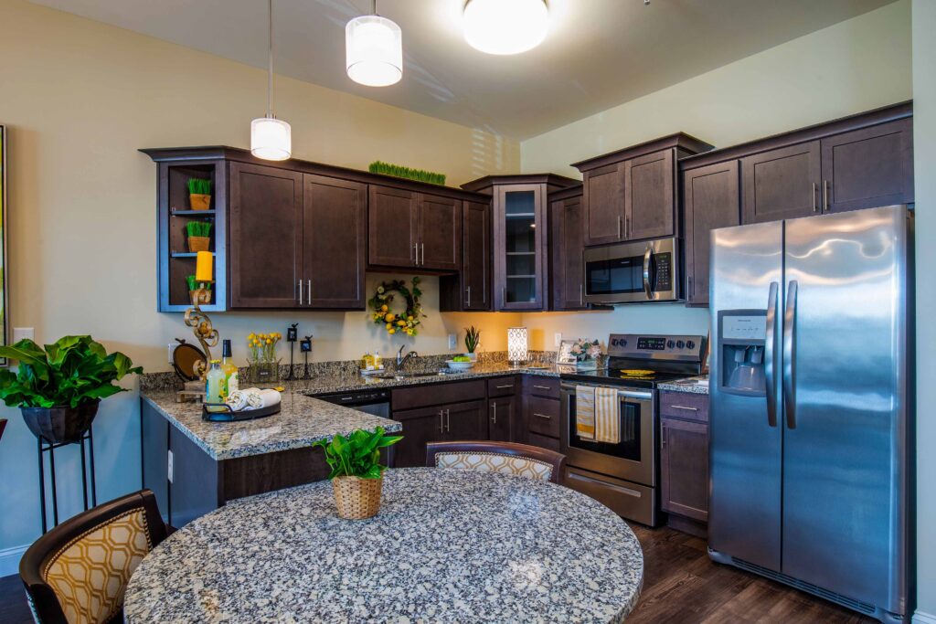 A kitchen with gray and white marbled counter tops and dining table. A potted plant is on top of the table with two brown, orange, and white-patterned chairs around it. The kitchen has brown cabinets, a dishwasher, an oven, a microwave, and a fridge.